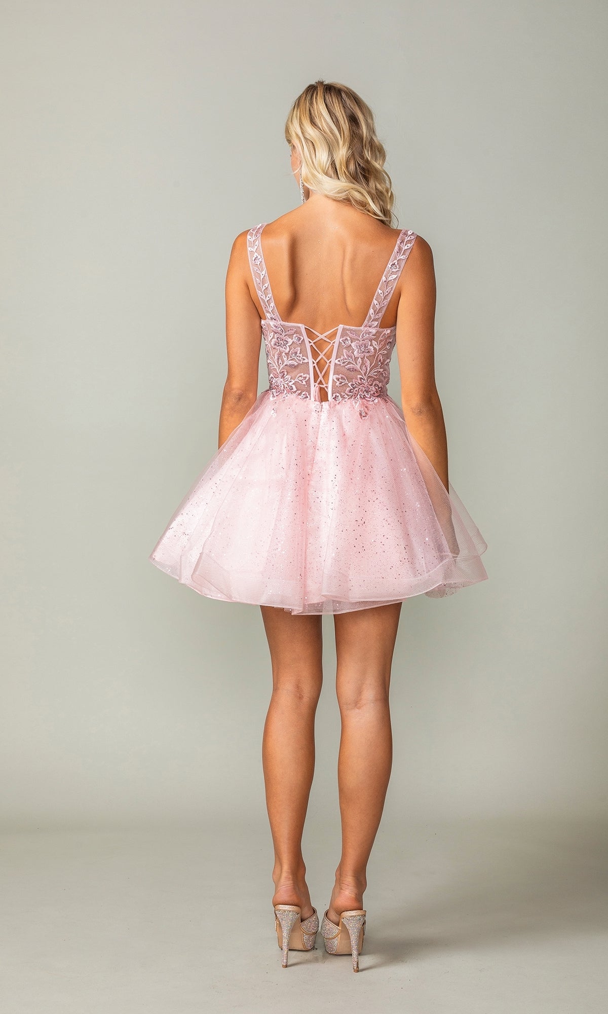 A woman faces away, wearing the Embroidered-Bodice Short Lace-Up Hoco Dress 3390 in light pink, featuring a lace-up back and tulle skirt, styled with high heels.